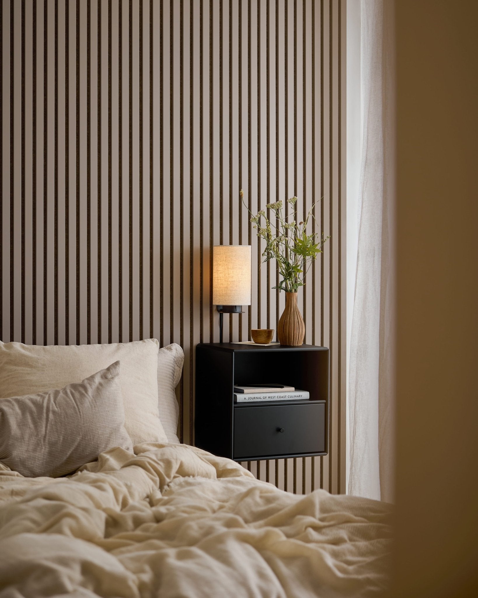 A bed with cream bedding beside a black nightstand, in front of an Akupanel wall in Dark Cashmere.