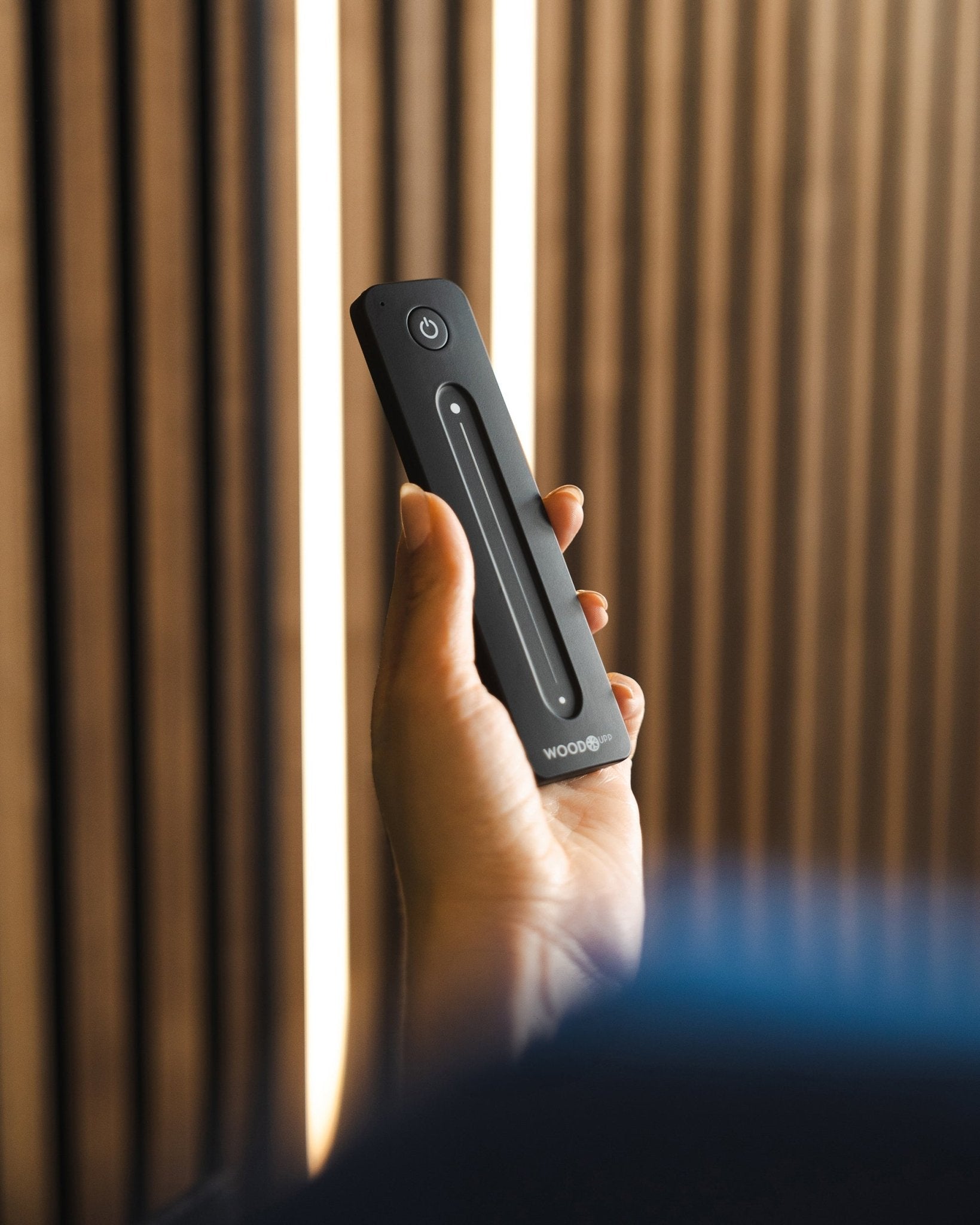 A hand holds a black WoodUpp Lumi LED Striplight remote with power button, positioned in front of WoodUpp acoustic slatwall panels; Philips Hue compatible.