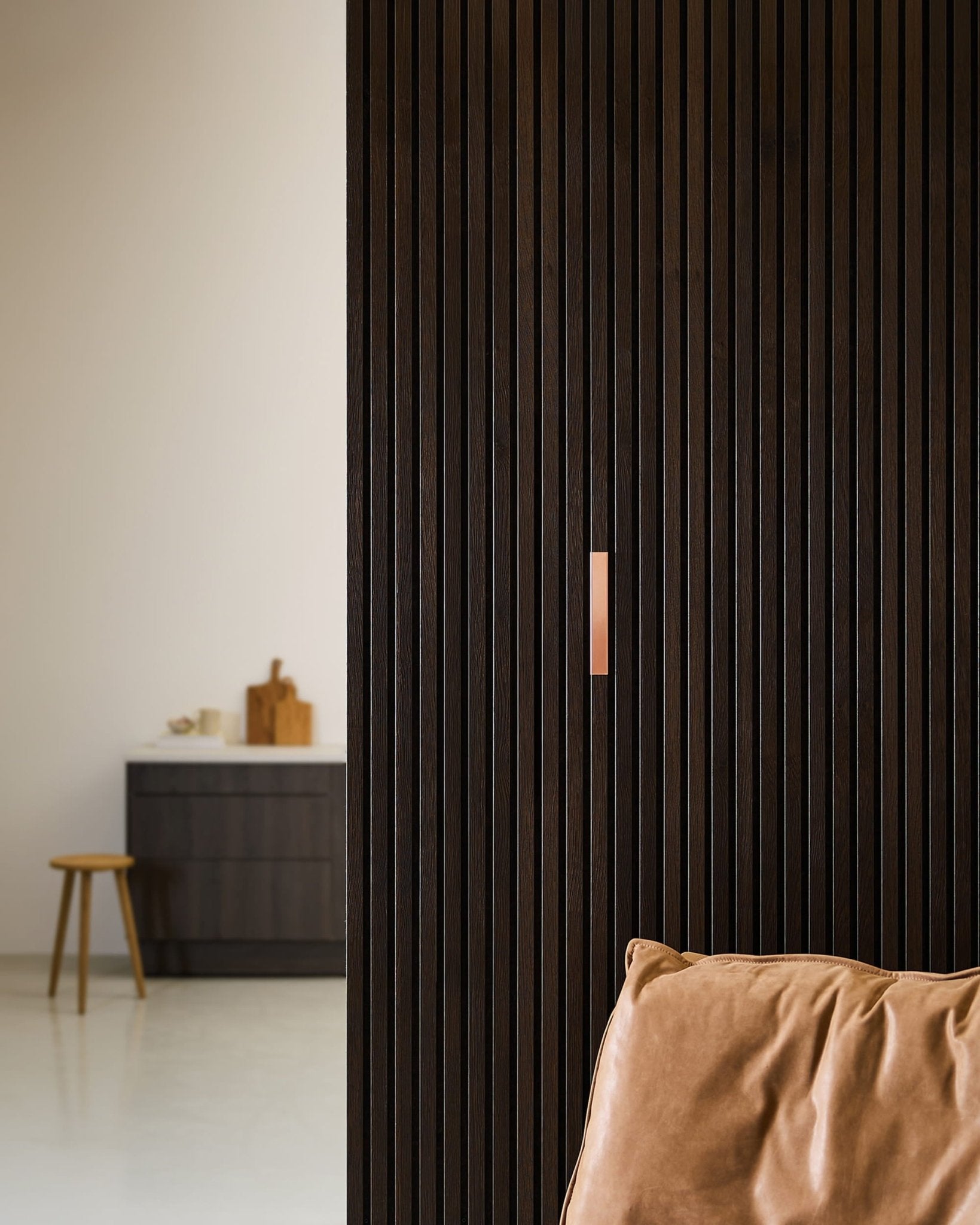 Minimalist interior with a brown leather chair, WoodUpp Liva | Cover / Copper 20 cm acoustic panels on the wall, and a stool by a counter with cutting boards.