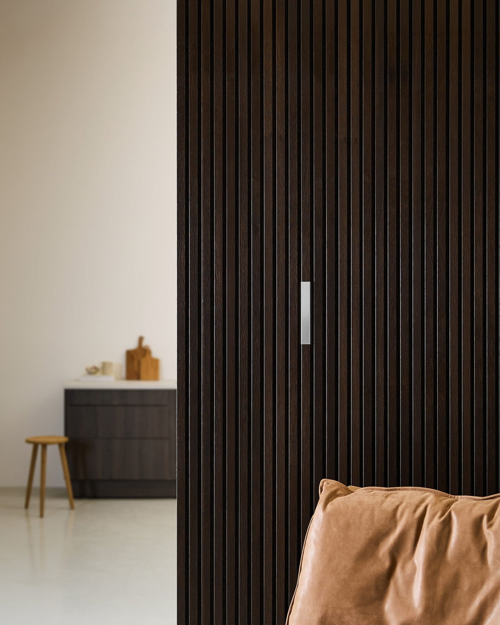 Brown leather chair beside a dark wood acoustic panel wall with WoodUpp Liva Cover in polished steel, adjacent to a minimalist kitchen and stool.