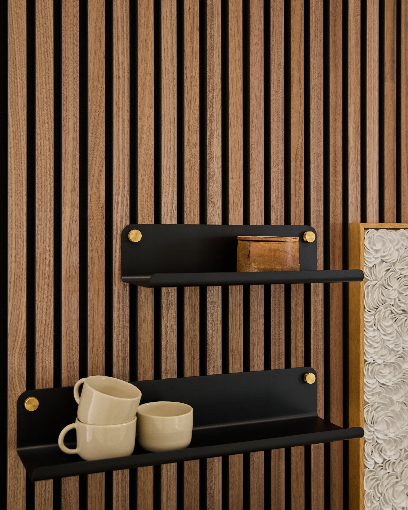 Two black Norr | Wide Shelves by WoodUpp (35.9 cm) with gold brackets mounted on an Akupanel acoustic wood panel; lower shelf holds 3 beige mugs, upper has a small wooden box.