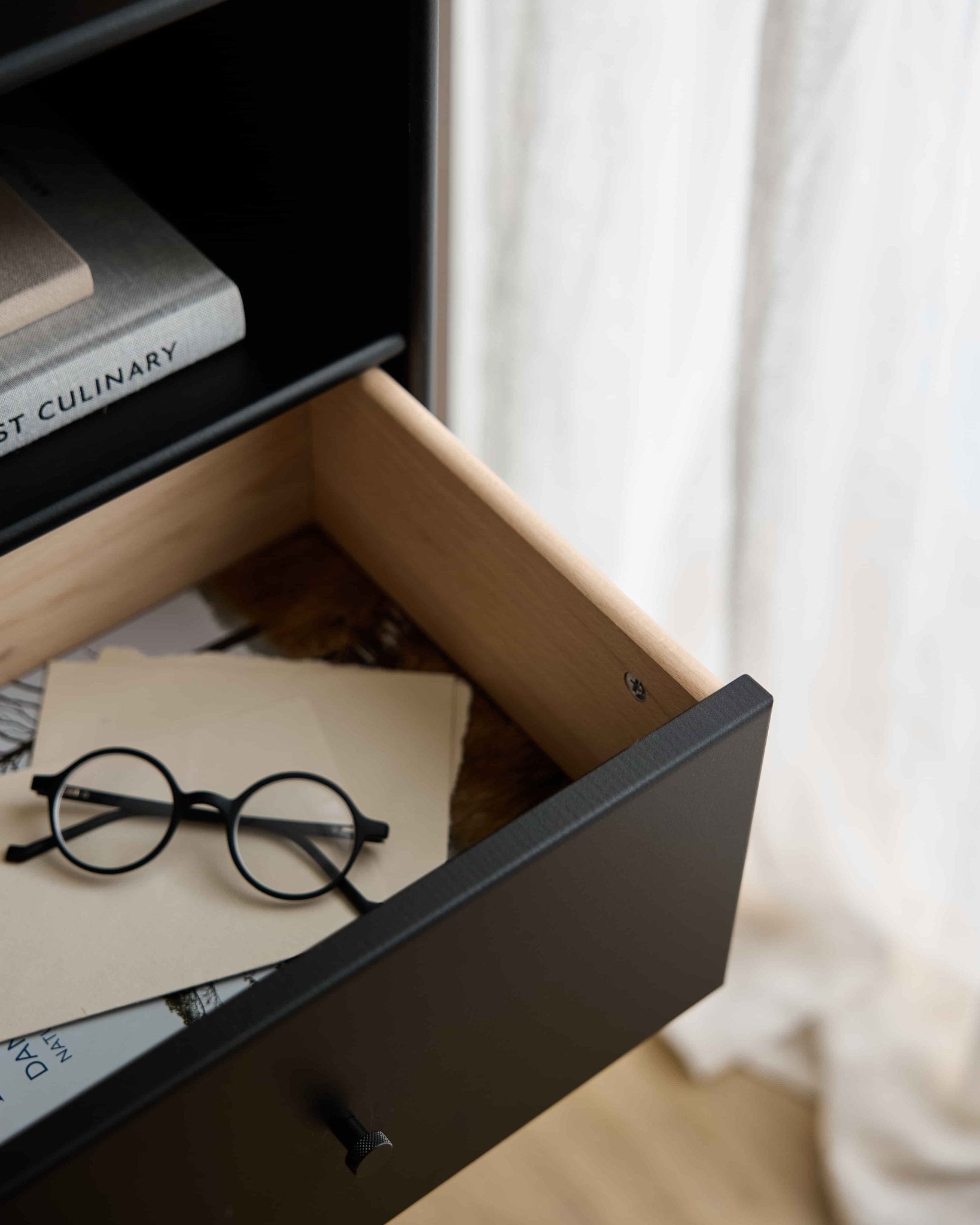 Eine runde schwarze Brille und Papiere in einer offenen WoodUpp Arka | Bücherregal-Schublade / Schwarz, unter Büchern, wobei transparente Vorhänge das Hintergrundlicht sanft streuen.