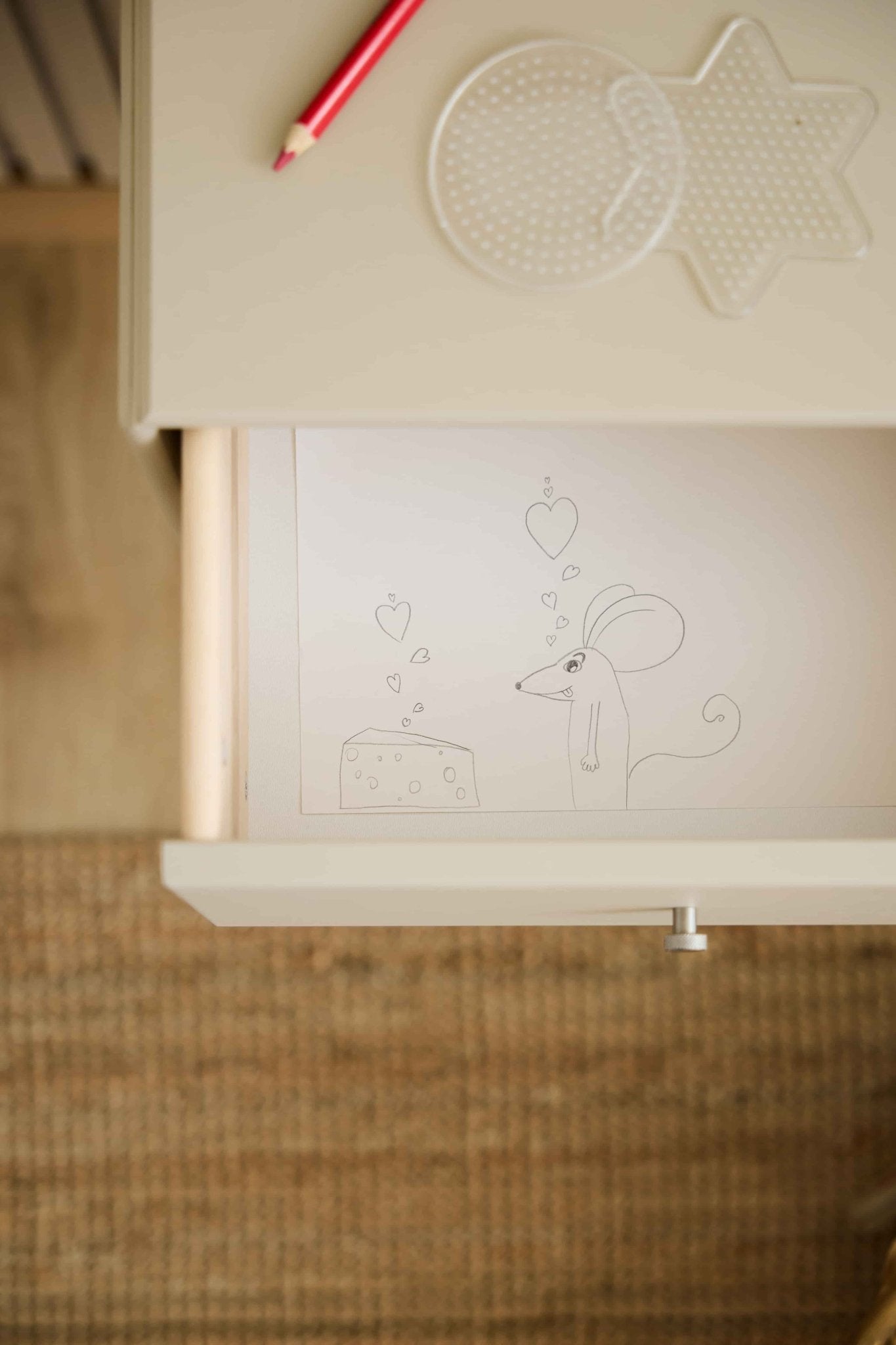 A mouse and cheese with hearts motif decorates a WoodUpp Arka bookcase drawer in beige; a red pencil and pegboards rest on the matching tabletop above.