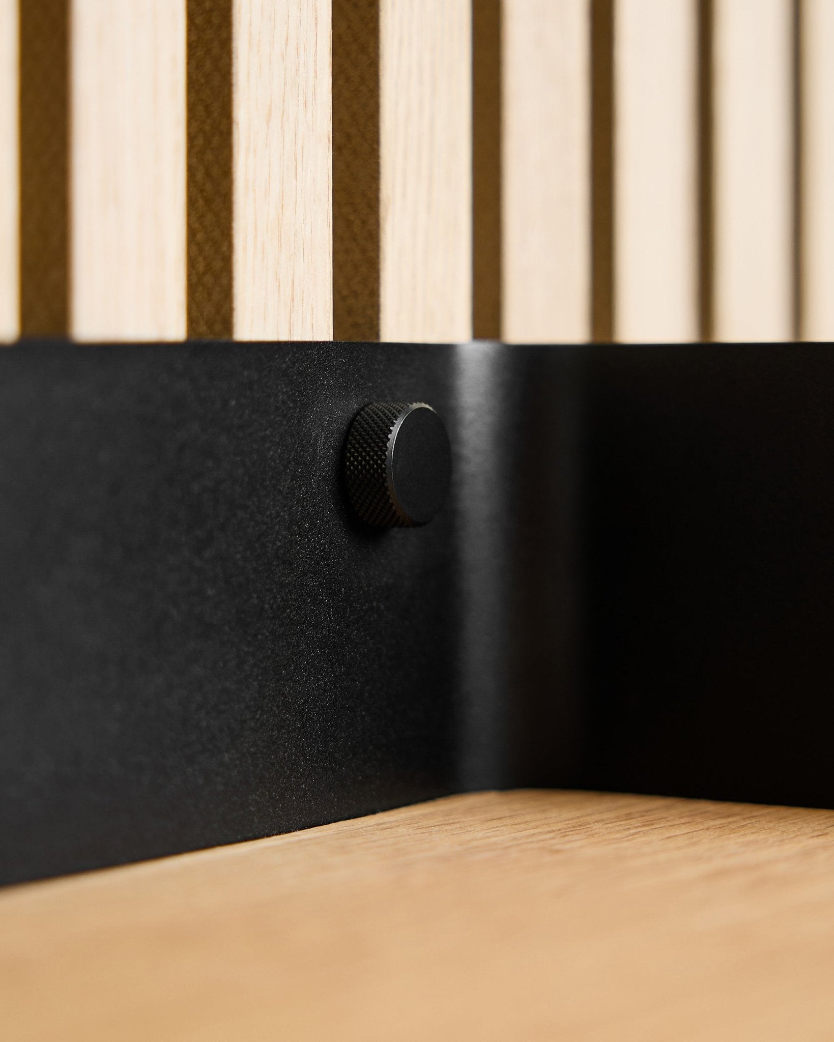 Close-up of two black WoodUpp Finger screws (M5x10 mm, 20x8.8) with brass screws, placed on a Nordic wood acoustic slat wall and light wood surface.