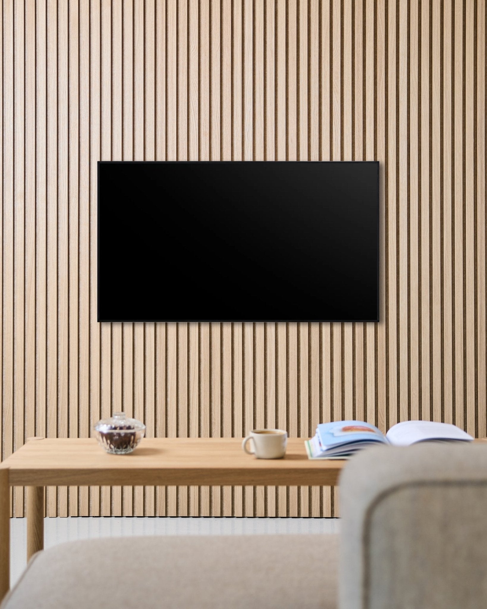 A Fjell TV Mount (small) by WoodUpp secures a flat-screen to a Nordic Akupanel acoustic wood slat wall, above a table with cup, bowl, and open magazine beside a beige sofa.