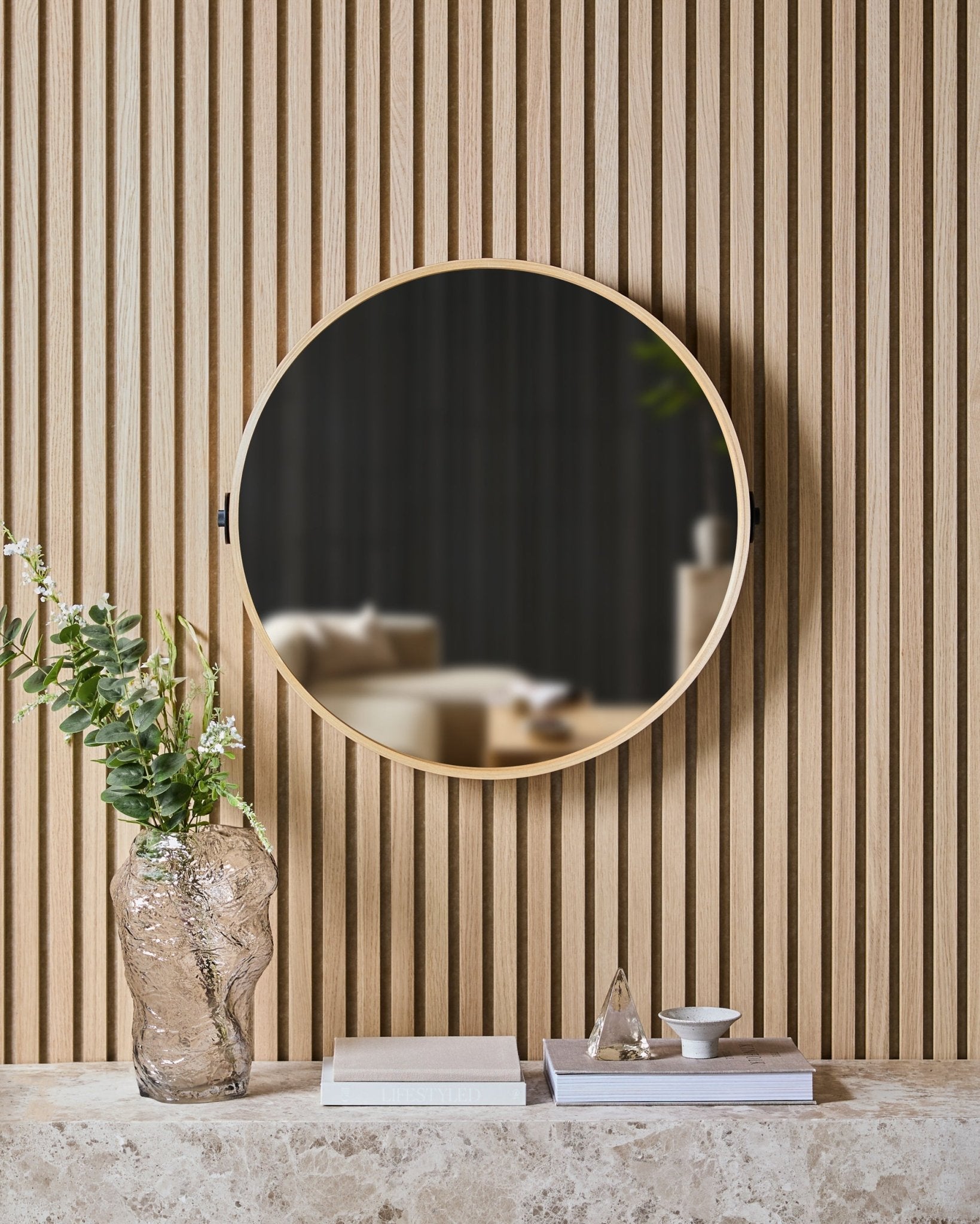The Linn | Mirror in Natural Oak by WoodUpp is mounted on a Nordic slatwall acoustic panel above a stone shelf with a glass vase, books, and decor items.