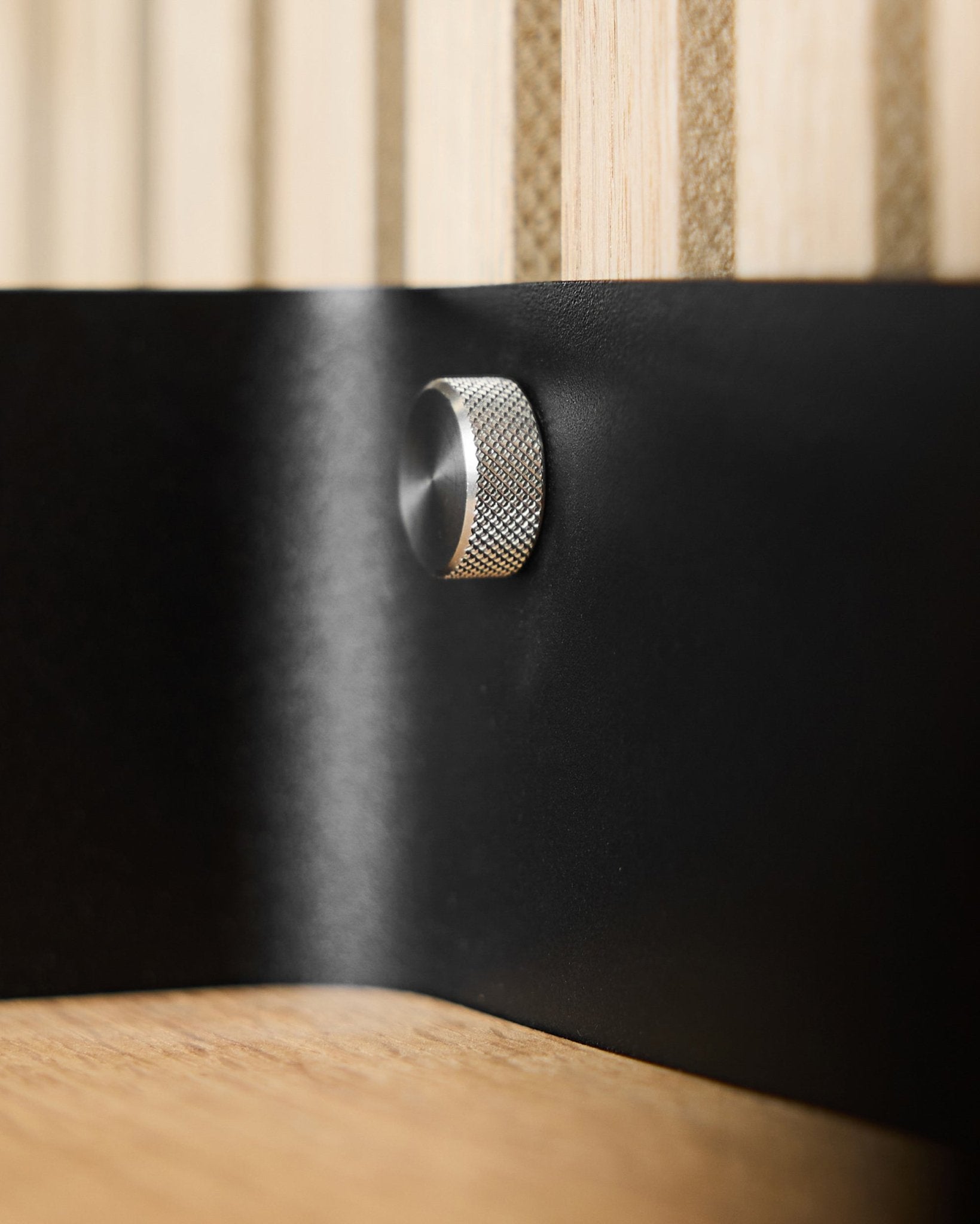 Close-up of two WoodUpp Finger screws (M5x10 mm, 20x8.8 stainless steel) on a black metal surface with Akupanel acoustic wood slats in the background.