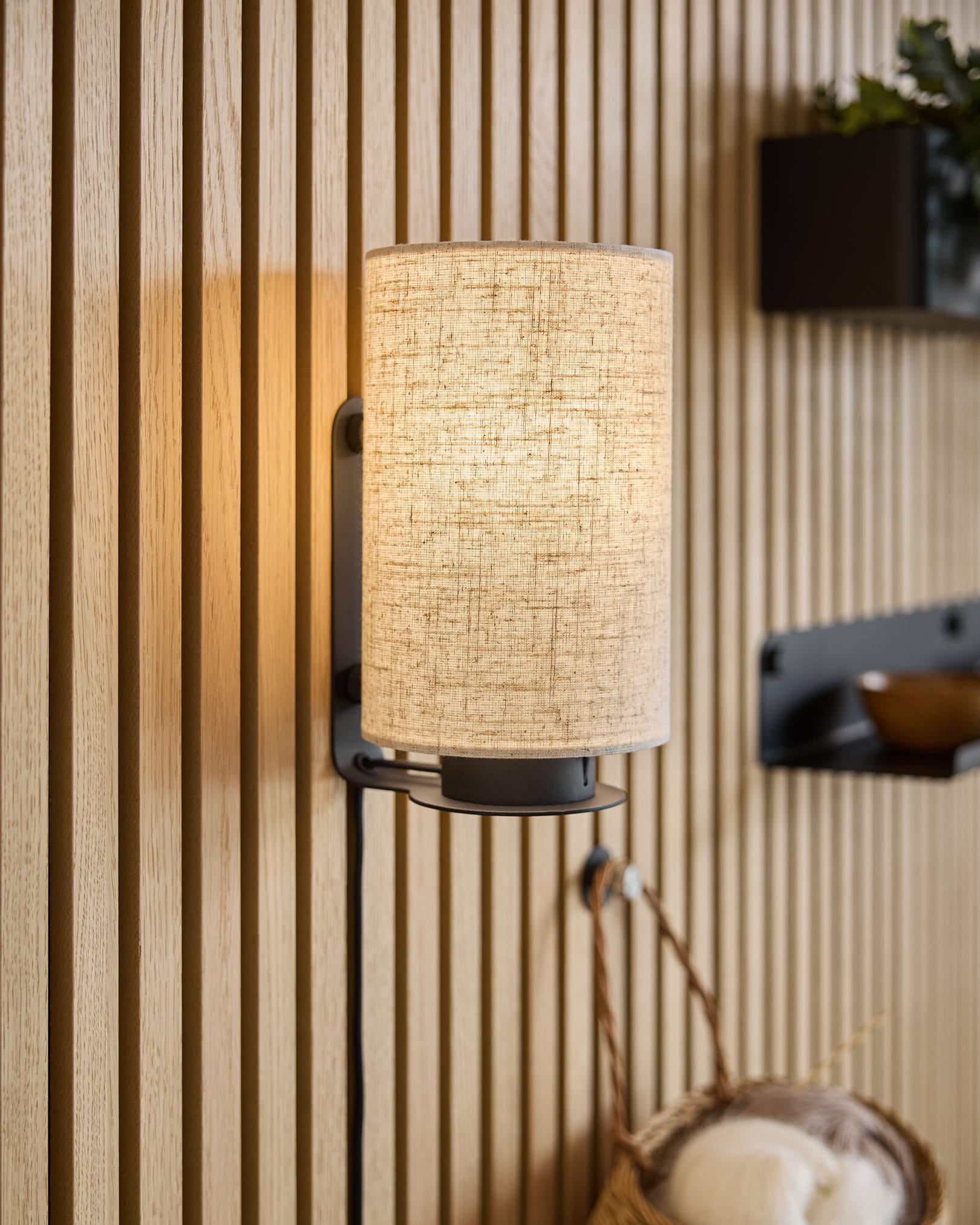 A modern wall sconce with a fabric shade casts warm light onto textured Akupanel acoustic wood panels, complemented by minimalist shelves and a woven basket.