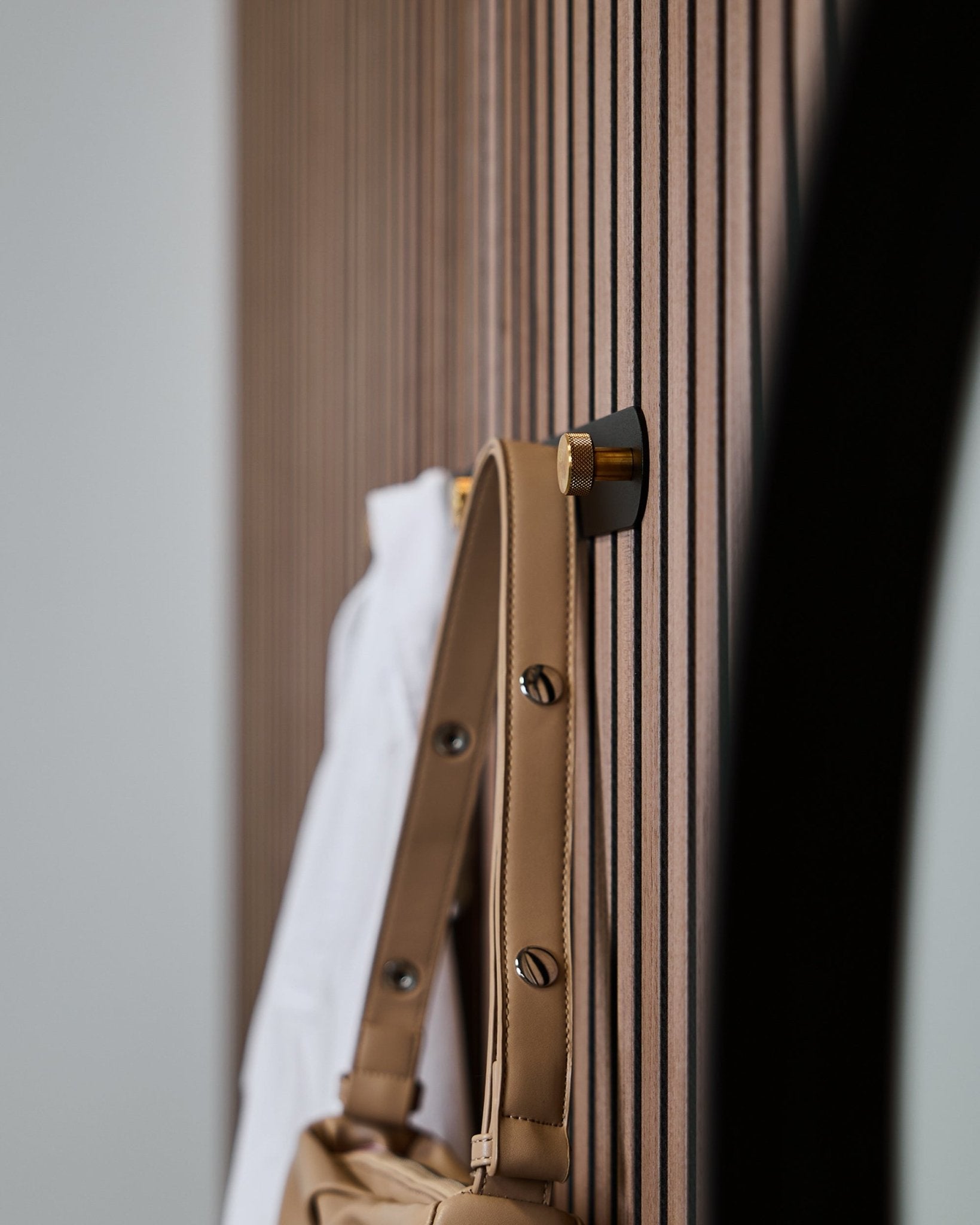 A tan bag and white shirt hang on a WoodUpp Norr Coat Hook Rack (Black/Brass, X5) mounted to a vertical Akupanel acoustic slatwall panel.