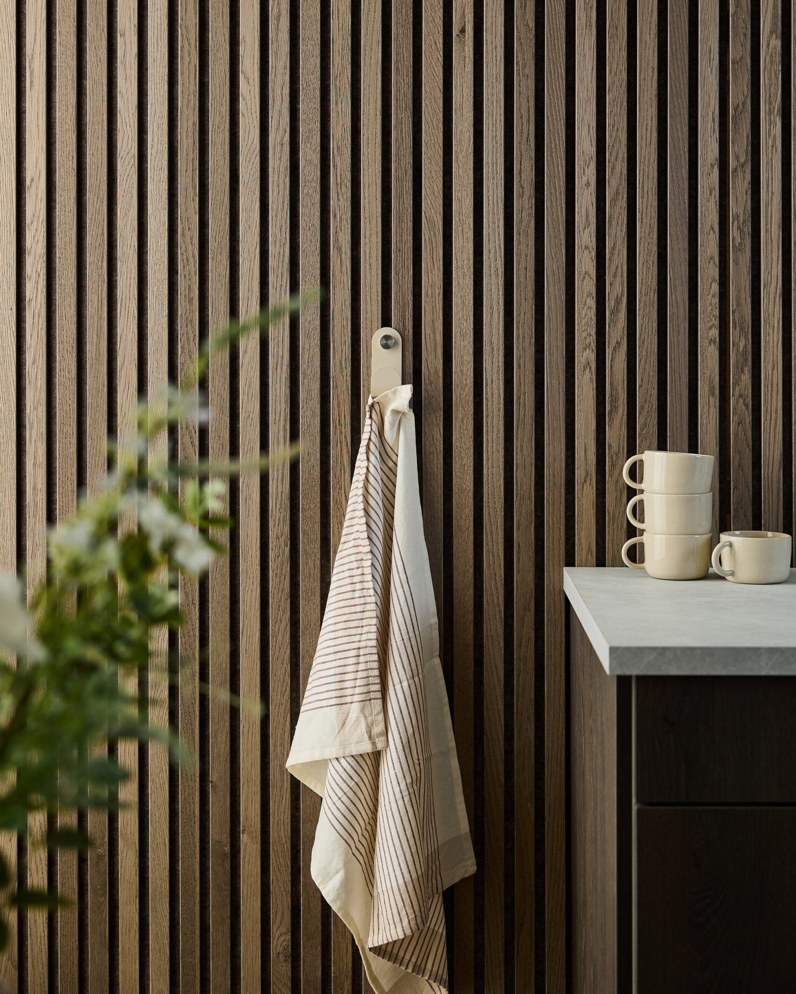 A striped towel hangs from a beige WoodUpp Norr Single Hanger mounted on a wood slat acoustic panel; stacked mugs and a plant are on the counter below.