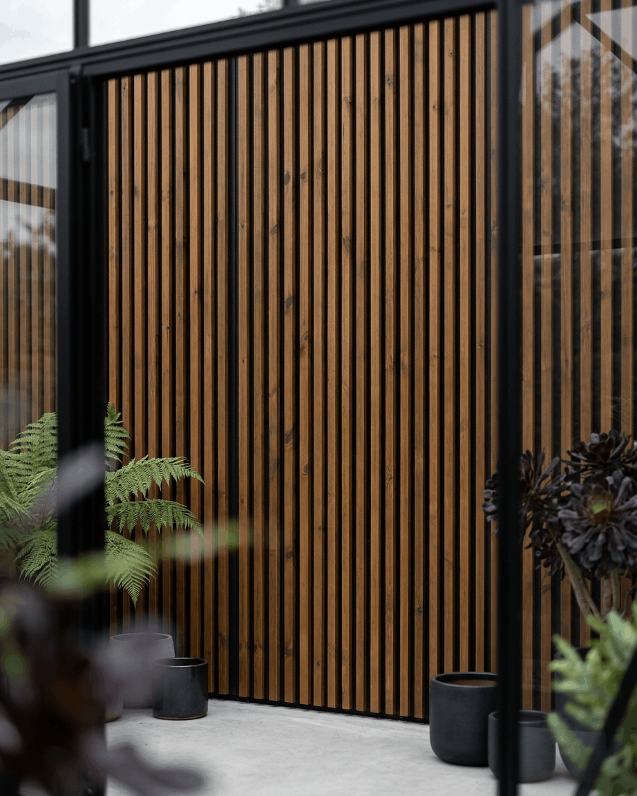 WoodUpp Aluwood outdoor facade panel in oiled brown paired with black planters and green plants, creating a minimalist Nordic patio setting.