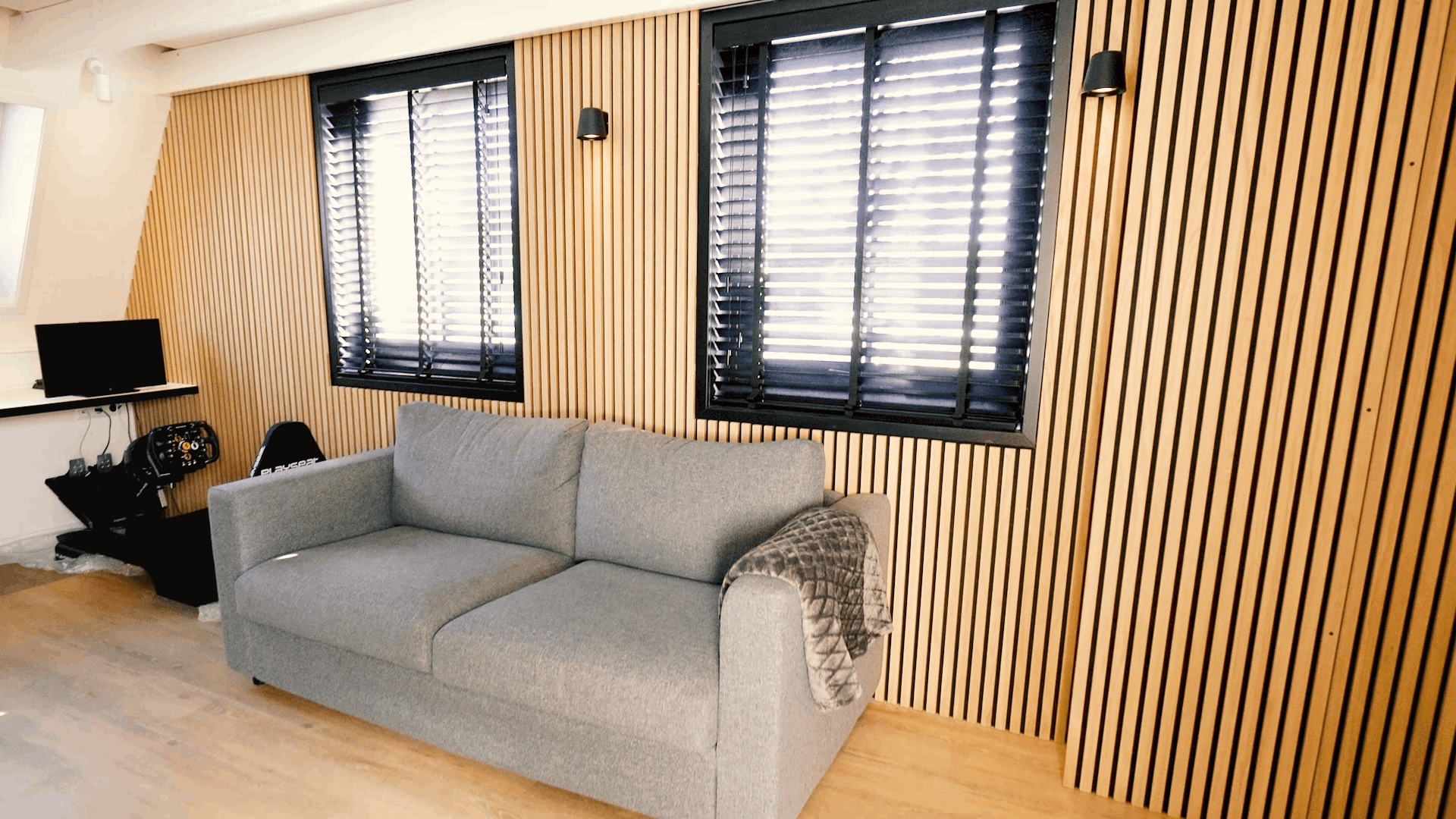 Gray sofa with patterned blanket placed in front of a Nordic Akupanel acoustic wood slat wall, flanked by two windows with black blinds and a compact desk setup.
