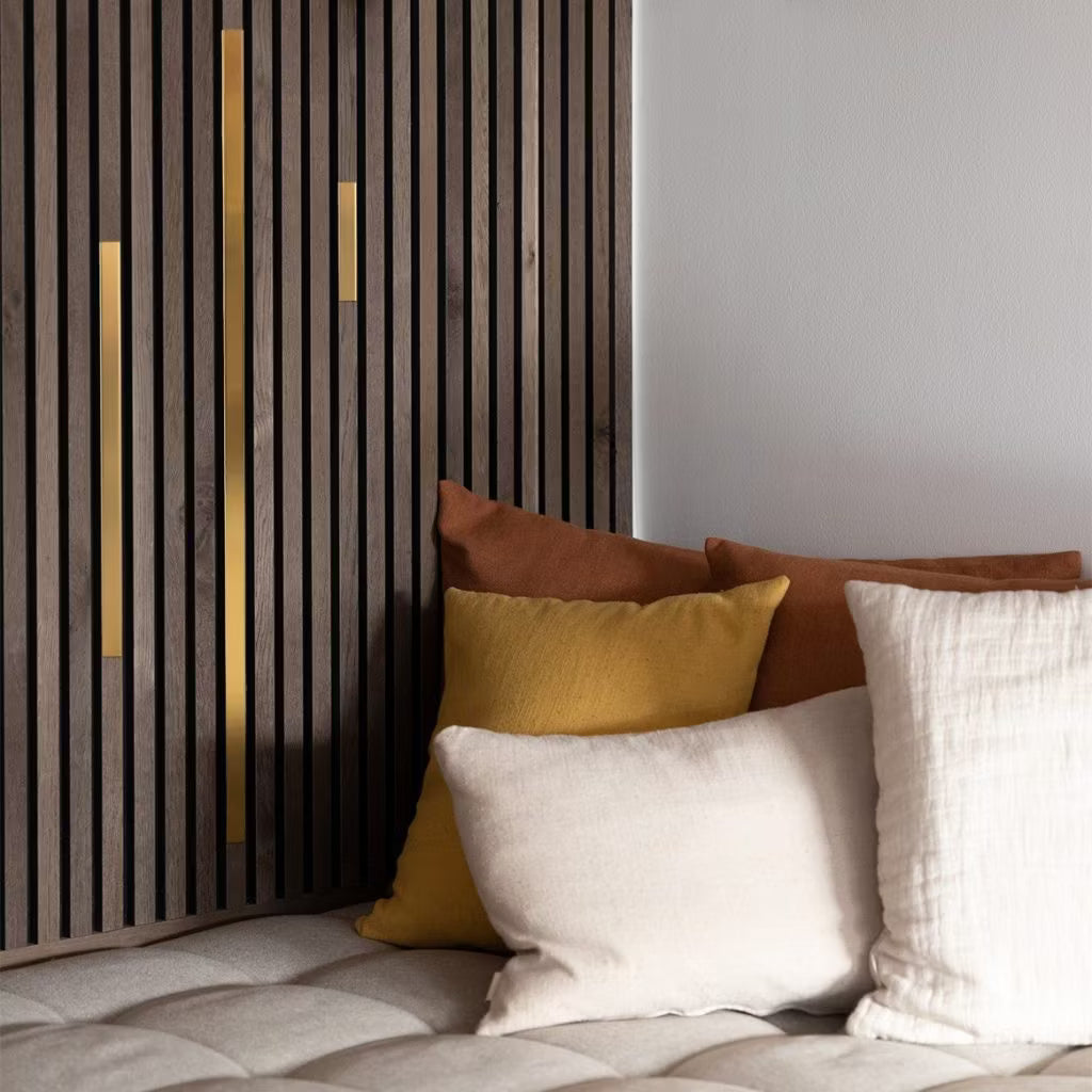 Cream, yellow, and brown cushions on a beige tufted sofa beside a modern acoustic slatwall panel with gold accents for a cozy, contemporary setting.