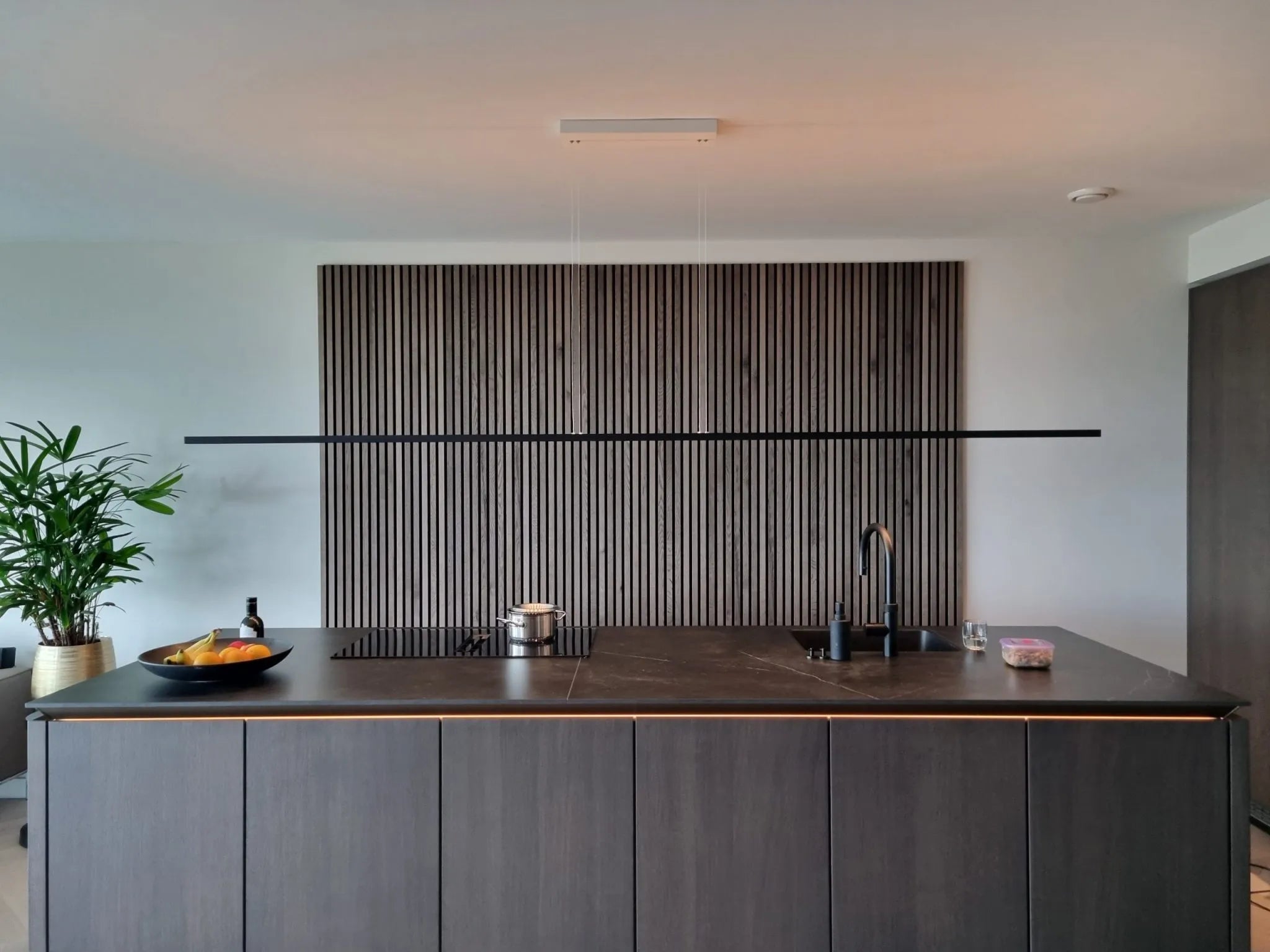 Modern kitchen with a dark island, built-in sink and stove, fruit bowl, and potted plant. Vertical Akupanel acoustic wood panels accent the wall behind the island.