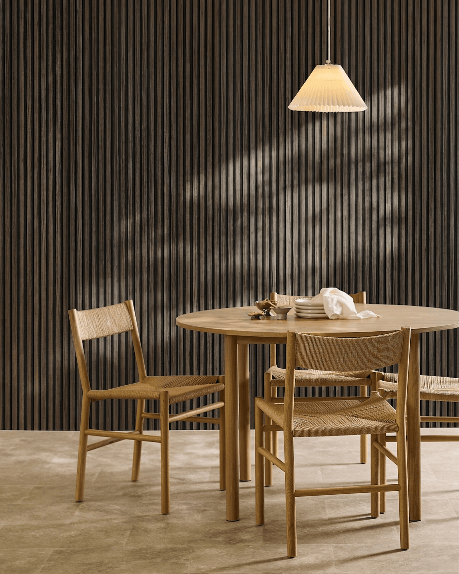 A round table with four woven chairs is arranged beside a WoodUpp Akupanel Black Oak acoustic panel, beneath a pleated pendant lamp.