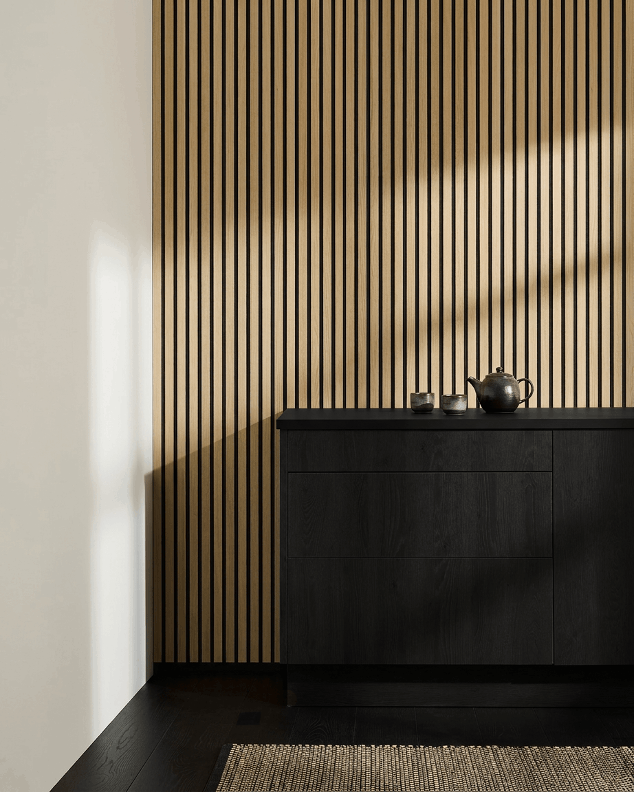 Minimalist Nordic kitchen featuring WoodUpp Akupanel acoustic panel in Classic Oak, black cabinet, teapot, cups, and woven rug on dark wood floor.