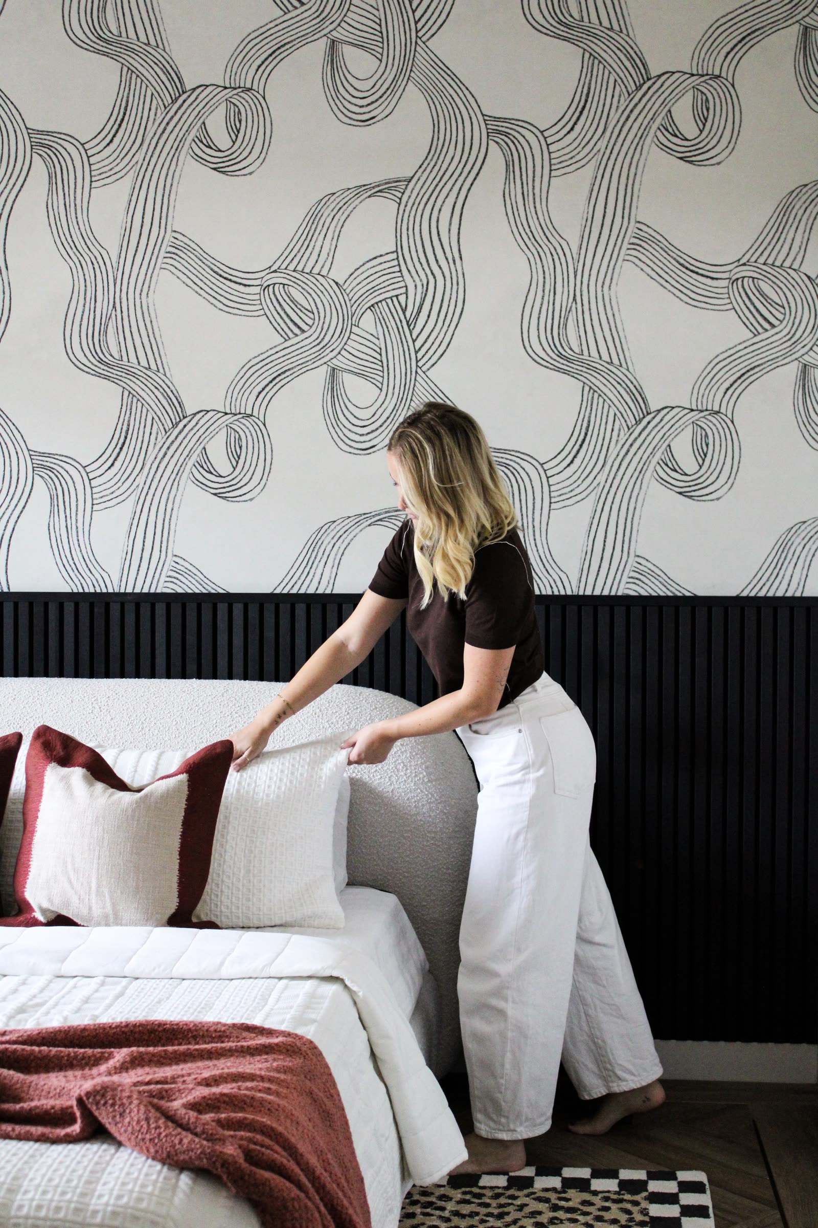 Woman dressing the bed against black Akupanel half-height panelling under graphic wallpaper