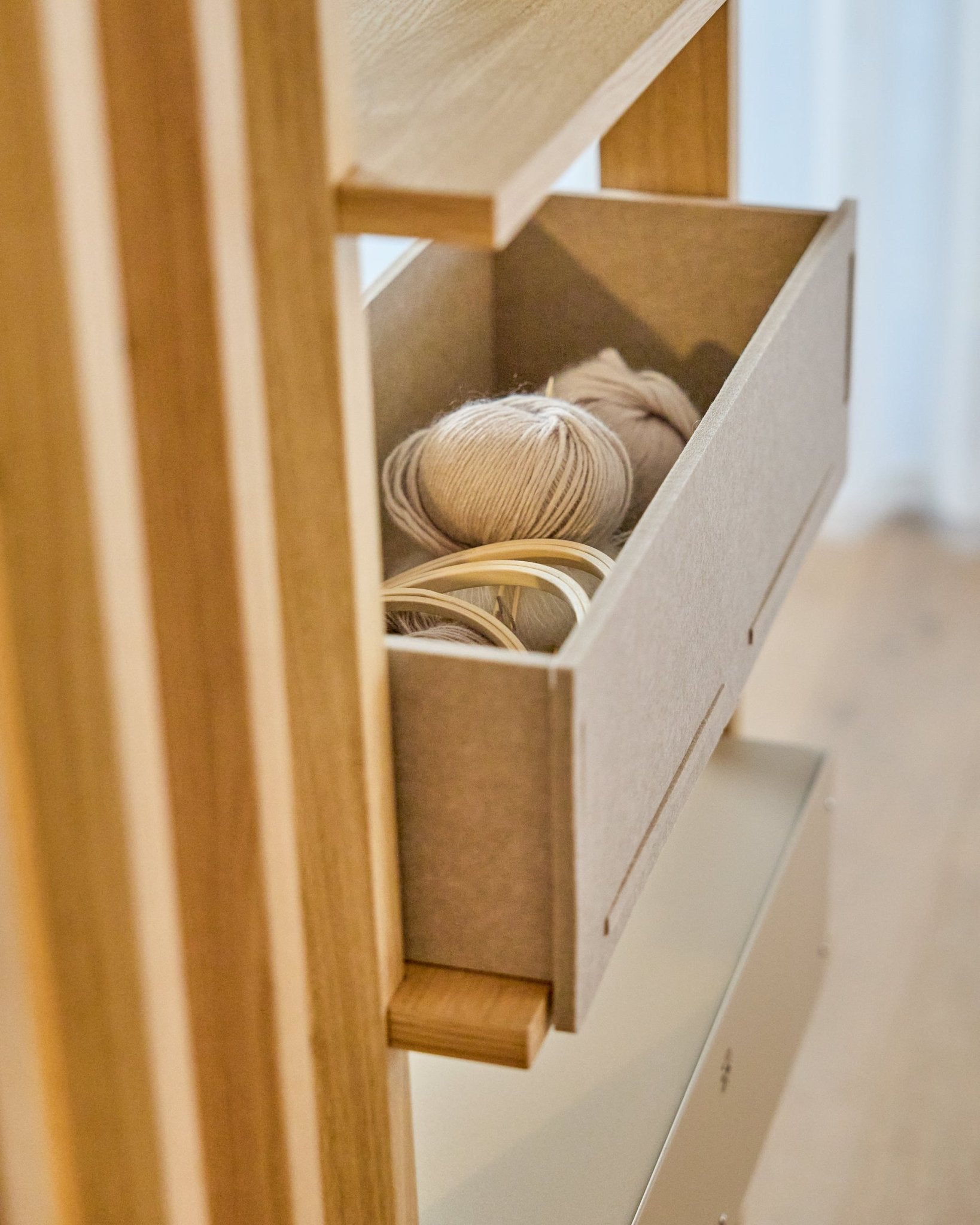 The WoodUpp Vigg Felt Box / BM2 in beige holds light yarn balls and wooden embroidery hoops on a wooden shelving unit, complementing Nordic acoustic panels.