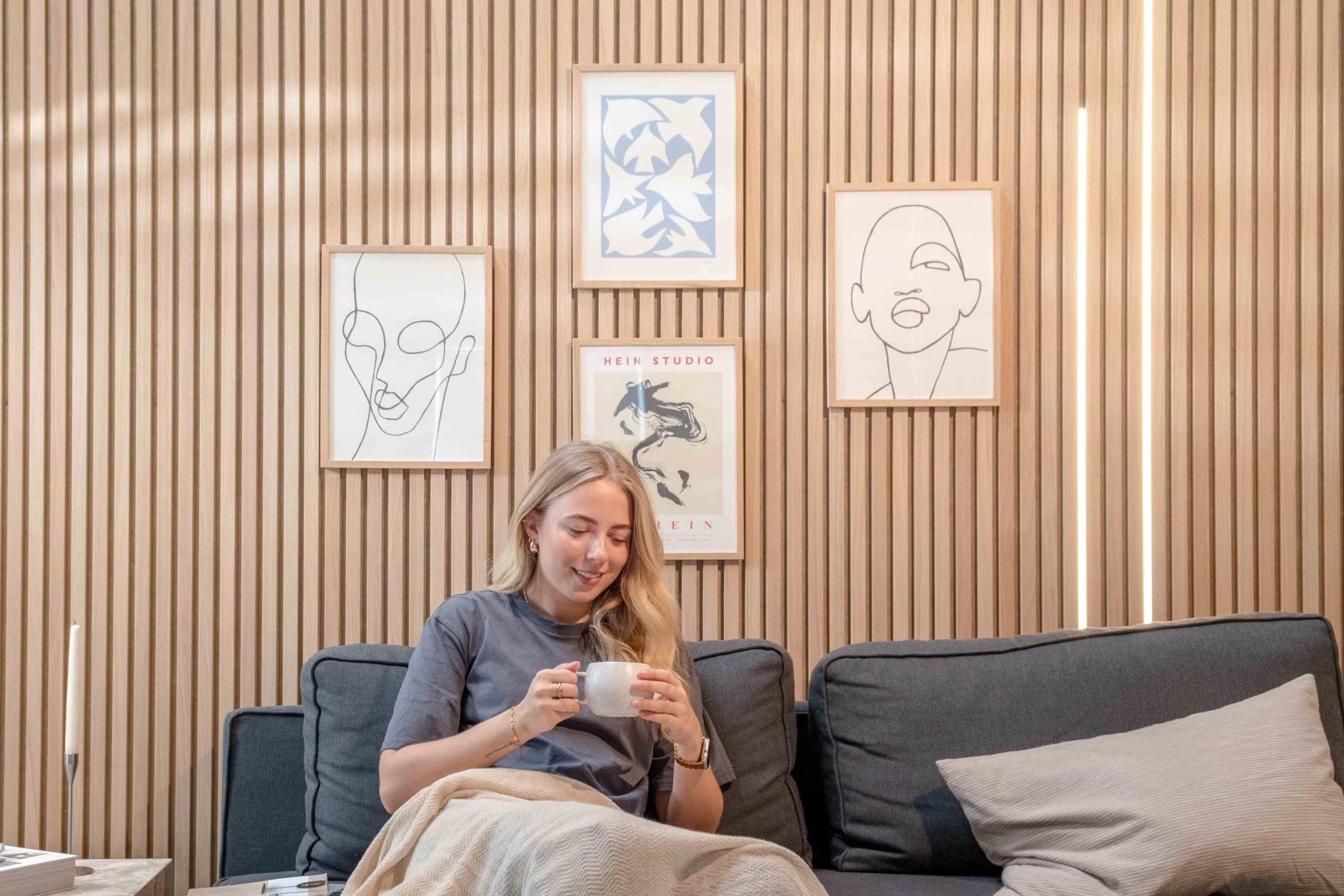 Woman relaxing on a sofa with a mug, smiling beneath minimalist line art and abstract decor mounted on textured Nordic acoustic slatwall panels.