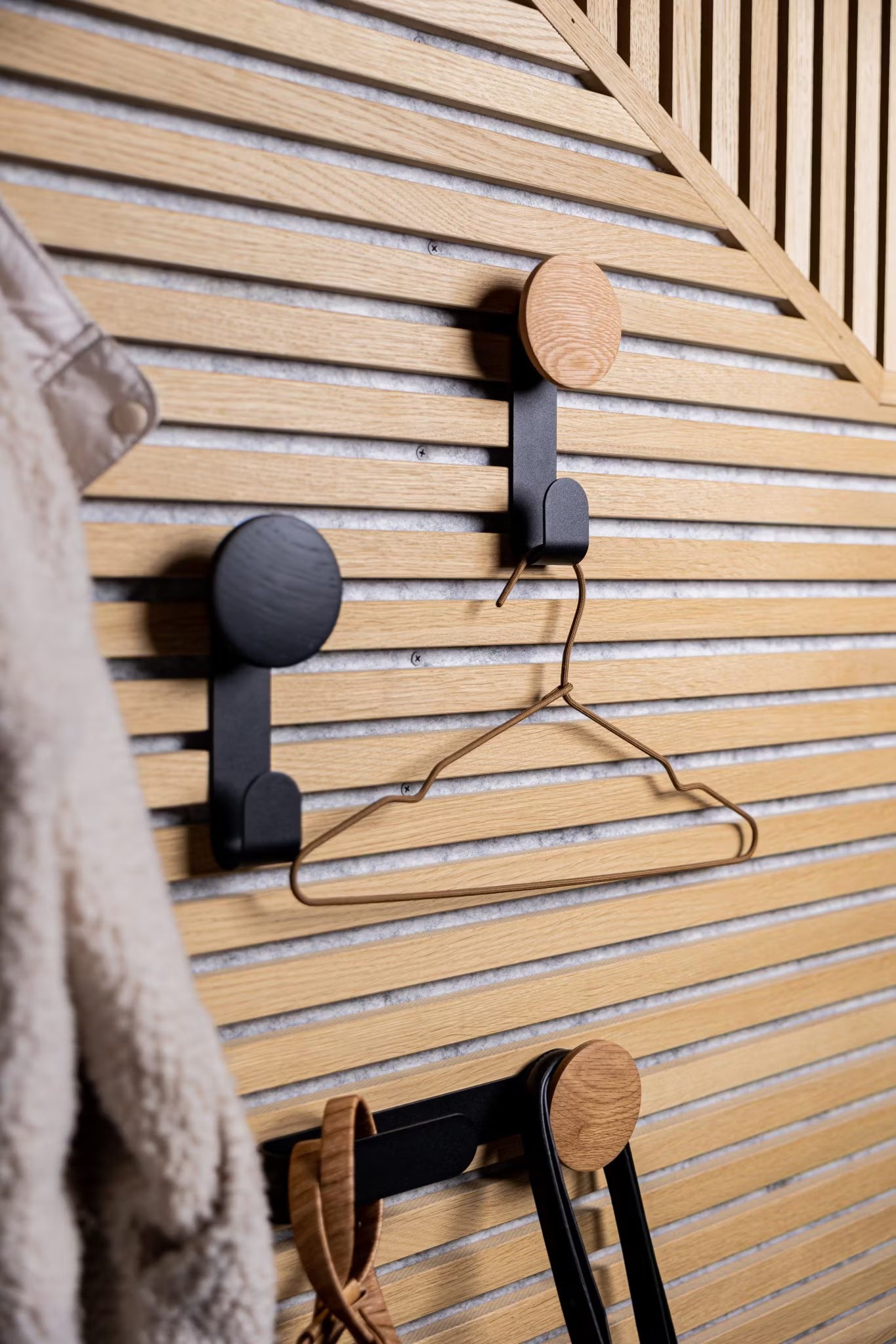 Three modern hooks mounted on a Nordic-style Akupanel acoustic wood slatwall hold a metal hanger and accessories, with a fuzzy coat partially visible on the left.
